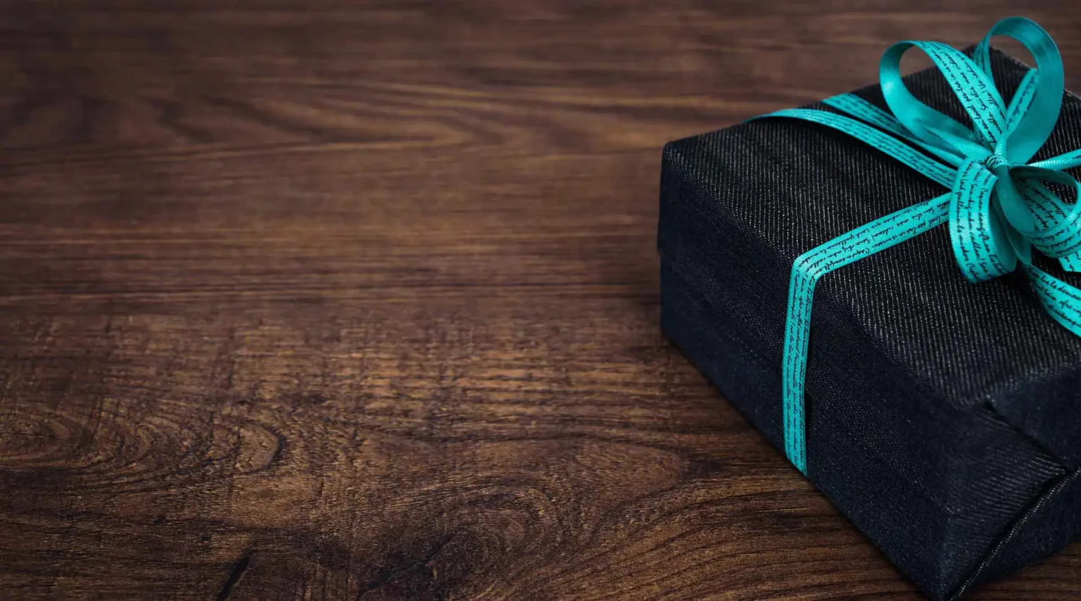 In the picture there is half of a small gift box with a black paper and a light blue ribbon on a wooden table.