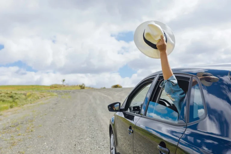 In the picture there is half of a blu car traveling on a dirt road. The windowof the back seat is open and from it the arm of a person appears holding a white hat.