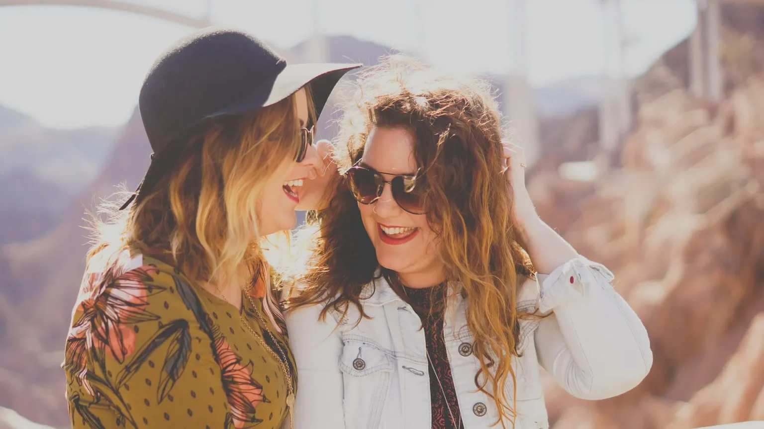 In the picture there are two girls laughing. They are outside and they are wearing sunglasses and, one of them, a large black hat.