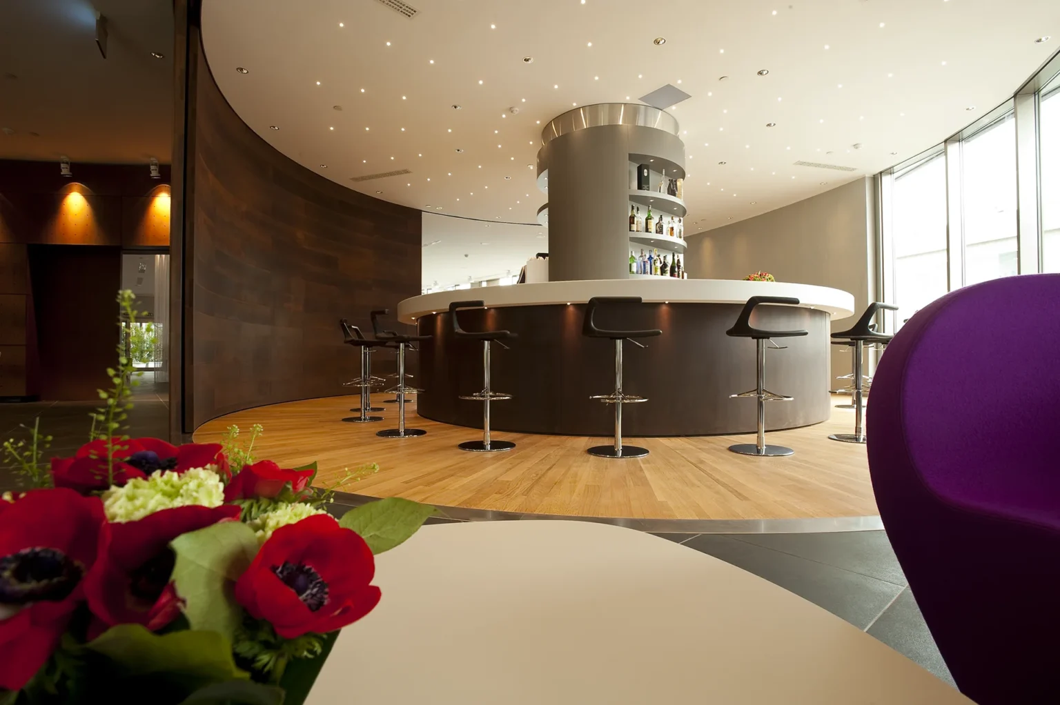 Nell'immagine, il Lounge Bar del The Hub Hotel 4 stelle a Milano: si vede il bancone del bar con degli sgabelli. Dietro il bancone, dei ripiani con delle bottiglie di alcolici. In primissimo piano, si intravede un vaso di fiori rossi e bianchi e, sulla destra, lo schienale di un divano viola.