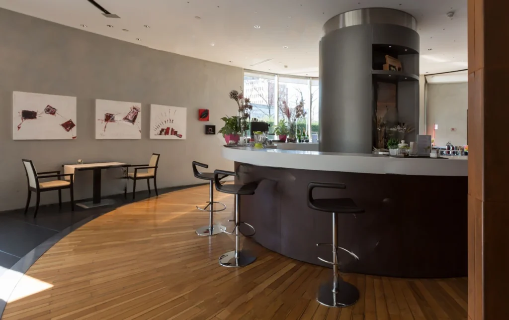 Nell'immagine, si vede il Lounge Bar del The Hub Hotel 4 stelle a Milano: in primo piano, il bancone del bar con tre sgabelli. Sulla destra, un tavolino con due sedie. Sullo sfondo, si intravede una vetrata.