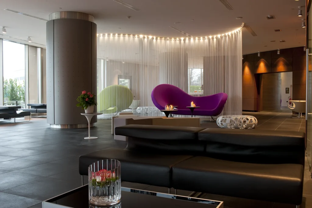 Nell'immagine si vede la sala Lounge del The Hub Hotel a Milano: in primo piano, un tavolino con un vaso di fiori e un divano nero. Sullo sfondo, un divano di colore viola acceso. Sulla sinistra, si intravedono altri divani neri e una vetrata.