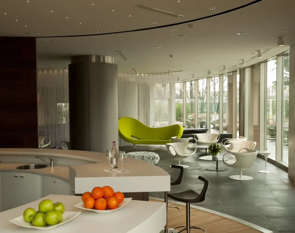 Nell'immagine si vede il Lounge Bar del The Hub Hotel a Milano: in primo piano, il bancone del bar con delle ceste di frutta e una bottiglia con due calici. Davanti al bancone, due sgabelli. Sullo sfondo, dei tavolini tondi con delle sedie e un divano.