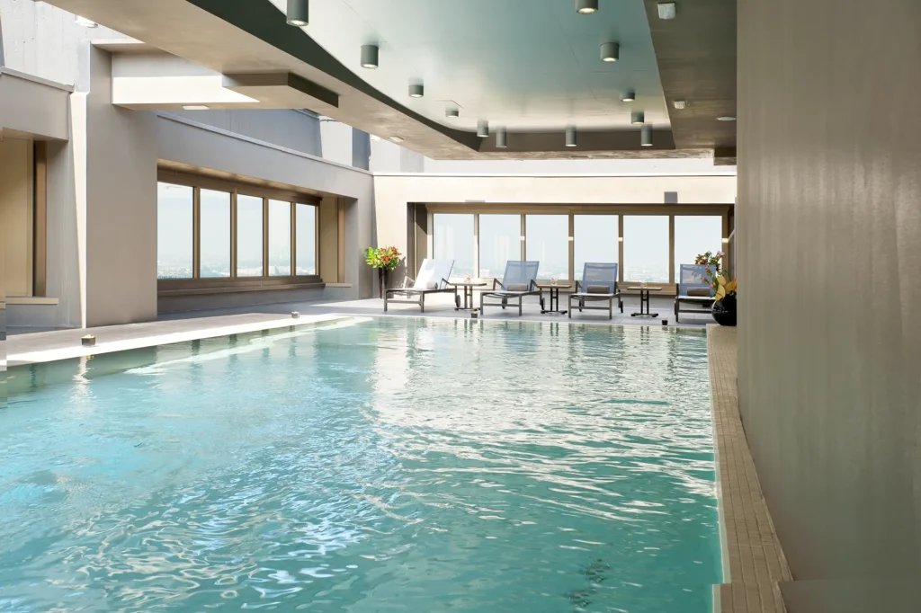 Nell'immagine si vede la piscina del The Hub Hotel con SPA a Milano: in primo piano, l'acqua della piscina e, sullo sfondo, quattro lettini. Tutto intorno ci sono ampie vetrate da cui entra la luce.
