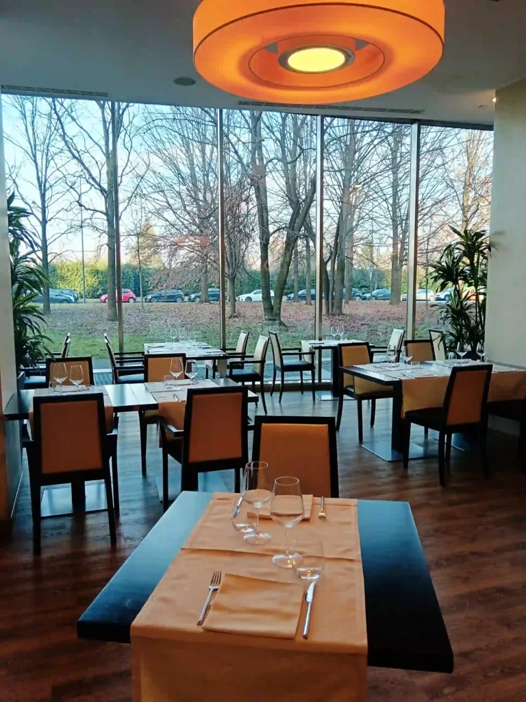Nell'immagine, una sala del ristorante del The Hub Hotel a Milano: in primo piano, si vede un tavolo apparecchiato e, di fianco, altri tavoli apparecchiati. Sullo sfondo, un'ampia vetrata e due piante.