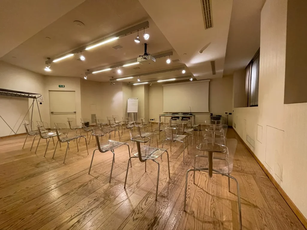 Nell'immagine, la sala meeting del The Hub Hotel: si vedono delle sedie disposte in file e, di fronte, lo schermo spento di un proiettore e una scrivania con due sedie. Sulla sinistra, una lavagna a fogli mobili.