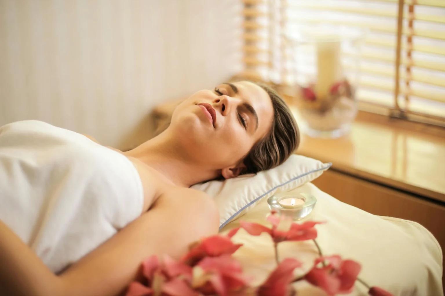 In the picture there is a girl covered with a towel relaxing on a massages bed. Next to her, a light candle and some flowers.