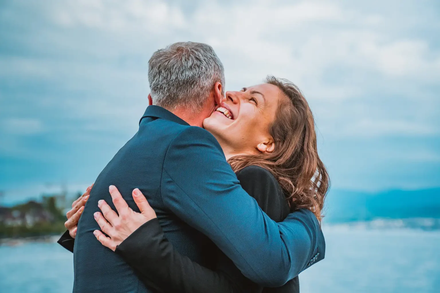 In the picture, a man and a woman hugging. The woman is smiling. In the background there is the sea.